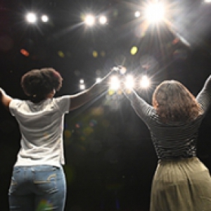 Two people standing side by side, lifting their hands together, facing bright lights