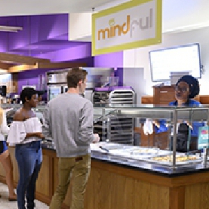 Students walking through dining hall area while other students stand at a cafeteria-style serving area