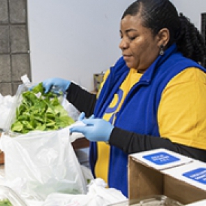 A woman in a yellow shirt and blue vest packs vegetables into a plastic bag