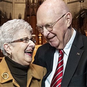 Roger Glunt in a black suit and red tie, holding hands and laughing with Lee Glunt, his wife, wearing a tan jacket. 