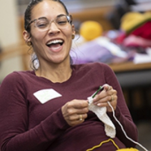 A woman in a maroon long sleeve shirt laughs while knitting