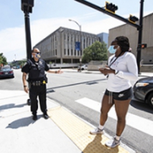 A police officer stands on a sidewalk, speaking to a person in a face mask in a white shirt and black shorts