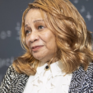 A woman in a black and white jacket with a white shirt underneath in front of a blackboard
