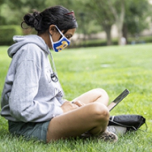 A student in a gray sweatshirt and a blue Pitt face mask works on a laptop in a field