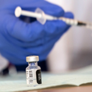 A vaccine bottle on a table with a blue gloved hand holding a syringe in the background