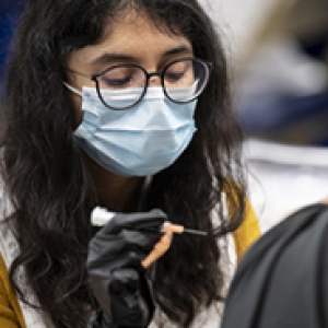 A person in glasses and a blue face mask holds a syringe in a gloved hand