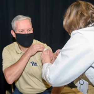 A man in a black face mask and a yellow Pitt shirt receives a shot