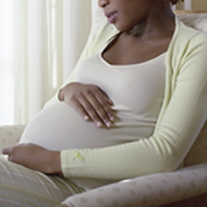 Pregnant woman sitting in a chair, holding her hands over her abdomen, wearing white shirt and light green cardigan.