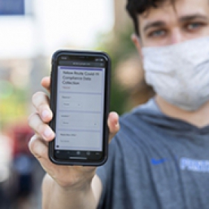 A person in a gray shirt and light face mask holds up a smartphone