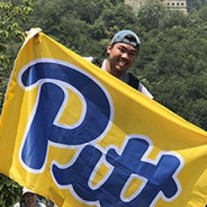 male student in a backwards hat holding a yellow and blue Pitt flag at the Great Wall of China