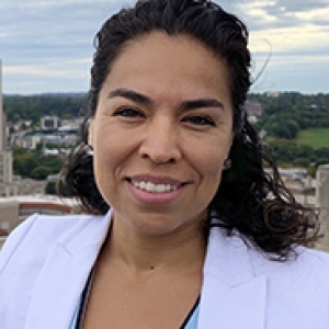 Gelsy Torres-Oviedo in white jacket over blue shirt, standing on a rooftop overlooking Pitt campus with Cathedral of Learning prominently in the background