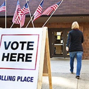 woman walking into a brick polling place building with a VOTE HERE sandwich board outside
