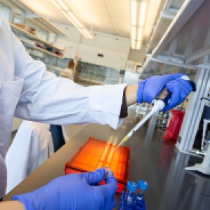 A person in a white lab coat and blue gloves uses a pipette