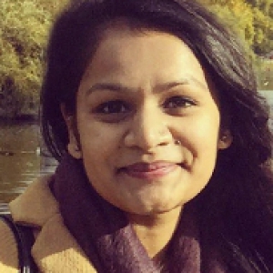 Patel in a tan coat in front of a body of water