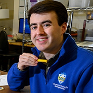 Tyler Bray (left) and Jacob Meadows (right) bioengineering seniors in Pitt’s Swanson School of Engineering, in blue Pitt jackets, sitting at a table with their Posture Protect vest on top.