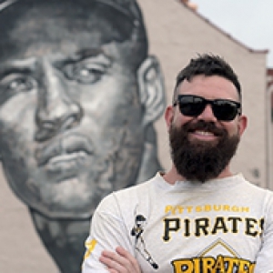 Pitt alumnus Jeremy M. Raymer in a PIttsburgh Pirates shirt standing in front of a mural he painted of Pirates baseball player Roberto Clemente