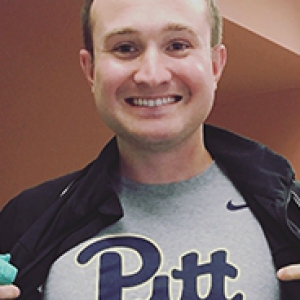 a young man holding some scrubs in one hand while pointing to the Pitt logo on his t-shirt