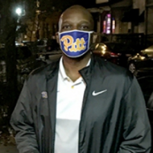 A man in a Pitt face mask and black jacket on a city street