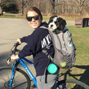 Katie Kniess with sunglasses on bicycle with Oliver, a black and white dog, in her backpack