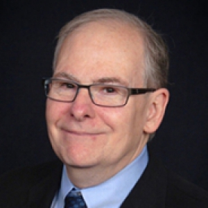 A man in glasses, a dark suit jacket and a light blue dress shirt