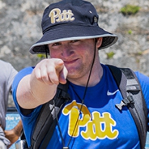 a student in a Pitt bucket hat and a Pitt t-shirt pointing at the camera and smiling