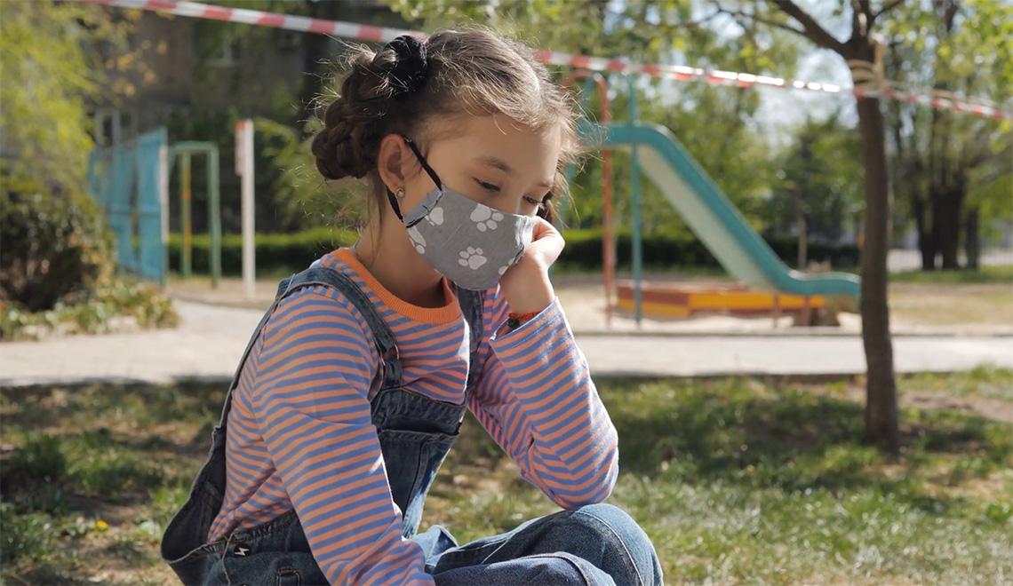 A girl wearing a face mask on a playground looks sad
