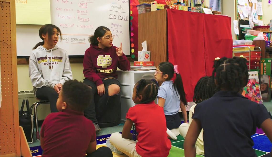 Teenagers sit in front of a class of elementary school students